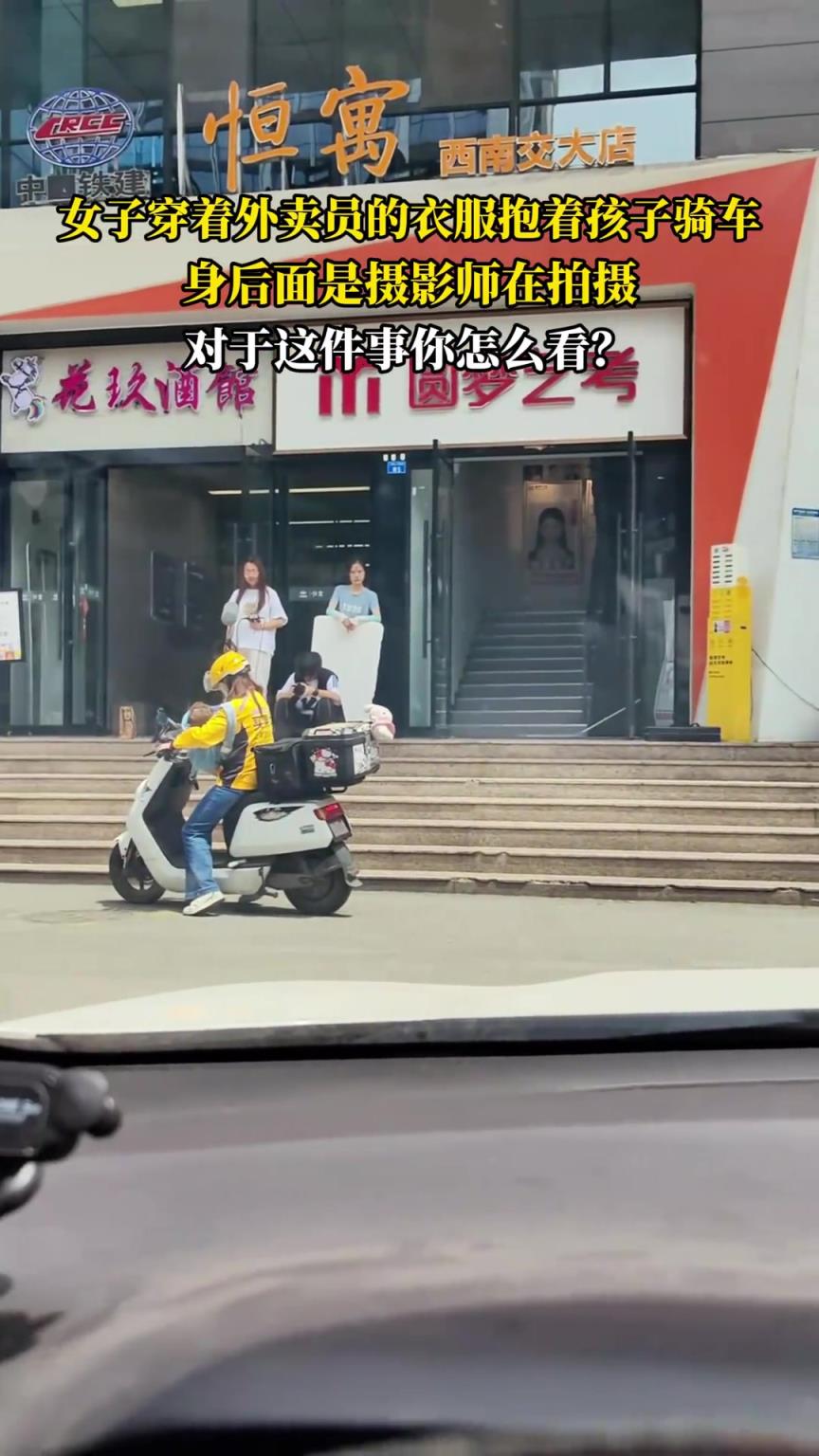 女子穿著外賣員的衣服抱著孩子騎車，身后面是攝影師在拍攝